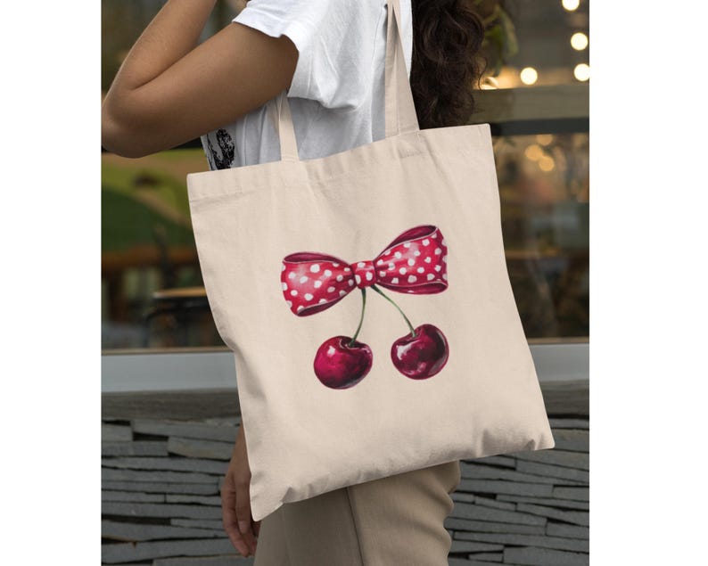 May include: A white canvas tote bag with a red and white polka dot bow and two red cherries. The cherries have stems and leaves.