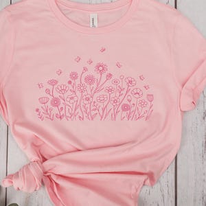 May include: A light pink t-shirt featuring a floral design in a darker shade of pink. The design includes various flowers and butterflies. The shirt has rolled-up sleeves and a tied knot at the bottom.