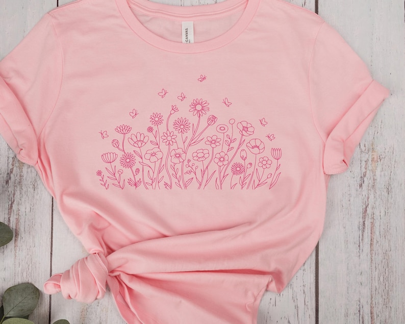 May include: A light pink t-shirt featuring a floral design in a darker shade of pink. The design includes various flowers and butterflies. The shirt has rolled-up sleeves and a tied knot at the bottom.