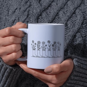 May include: White ceramic mug featuring a black line drawing of flowers growing out of rain boots. The design includes various floral types. The mug is held by a person wearing a gray sweater.