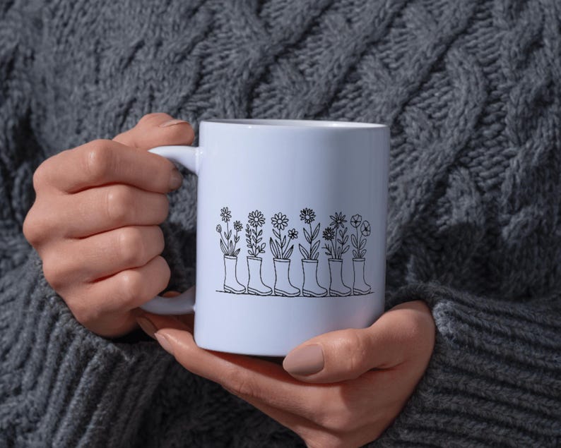 May include: White ceramic mug featuring a black line drawing of flowers growing out of rain boots. The design includes various floral types. The mug is held by a person wearing a gray sweater.