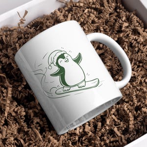 May include: White ceramic mug featuring a green illustration of a penguin snowboarding. The penguin wears a Santa hat and has its arms outstretched. The mug is inside a white box filled with brown crinkle paper.