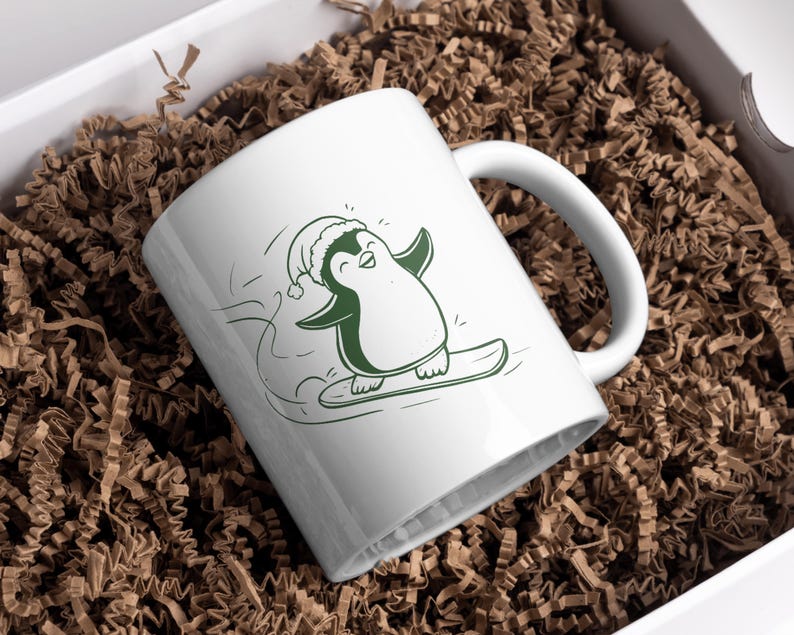May include: White ceramic mug featuring a green illustration of a penguin snowboarding. The penguin wears a Santa hat and has its arms outstretched. The mug is inside a white box filled with brown crinkle paper.