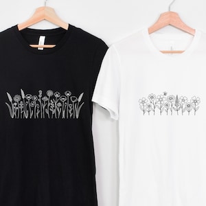 May include: Two t-shirts, one black and one white, each featuring a line drawing of wildflowers. The black shirt displays a variety of detailed floral illustrations, while the white shirt showcases a simpler design with a bee. Both shirts are on wooden hangers.