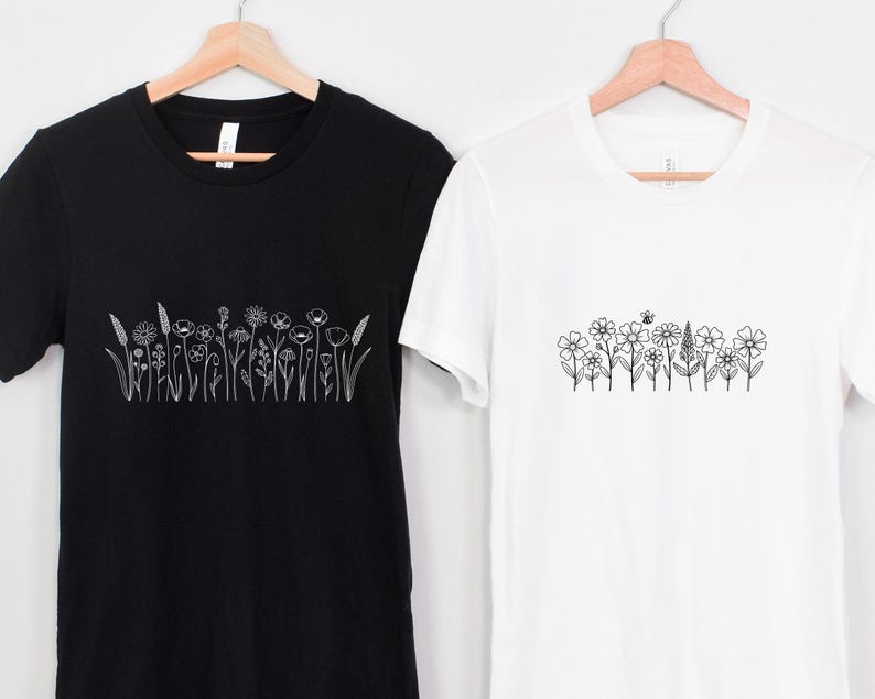 May include: Two t-shirts, one black and one white, each featuring a line drawing of wildflowers. The black shirt displays a variety of detailed floral illustrations, while the white shirt showcases a simpler design with a bee. Both shirts are on wooden hangers.