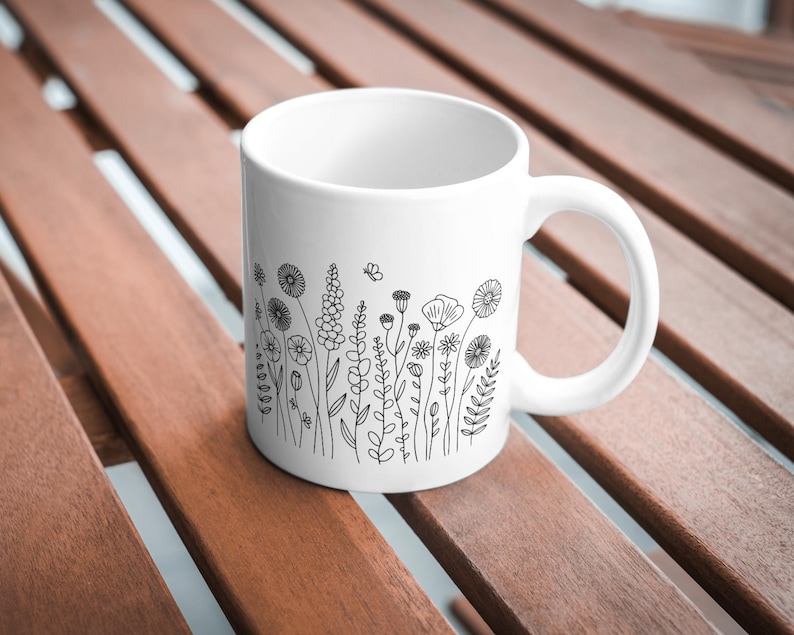 May include: White ceramic mug featuring a black line drawing of wildflowers and a butterfly. The mug has a curved handle and is set on a wooden surface. The floral design includes various flower shapes and leafy stems.
