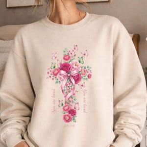 Puede incluir: Sudadera beige con un dise&ntilde;o de cruz floral. La cruz est&aacute; adornada con rosas rosas, hojas verdes y mariposas, con una cinta rosa. Las palabras "You are loved" y "John 3:16" est&aacute;n impresas en la sudadera.