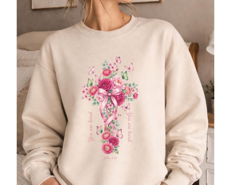 Puede incluir: Sudadera beige con un dise&ntilde;o de cruz floral. La cruz est&aacute; adornada con rosas rosas, hojas verdes y mariposas, con una cinta rosa. Las palabras "You are loved" y "John 3:16" est&aacute;n impresas en la sudadera.