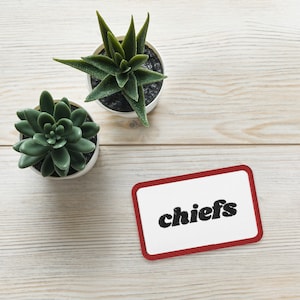 Pode incluir: Um emblema retangular bordado com um centro branco e uma borda vermelha, apresentando a palavra chiefs em letras minúsculas pretas. O emblema está posicionado perto de duas pequenas plantas suculentas verdes em vasos brancos sobre uma superfície de madeira clara.