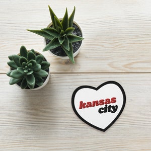 May include: A heart-shaped patch with the words "Kansas City" in red and black embroidery. Two small potted succulents are positioned above the patch on a light wood-grain surface.