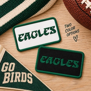 May include: Two rectangular embroidered patches featuring the word EAGLES in dark green lettering. One patch has a white background, and the other has a black background. The text TWO COLOR OPTIONS and a heart symbol are printed near the patches.