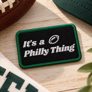 May include: A rectangular embroidered patch with a black background and a green border. The patch features white embroidered text that reads It's a Philly Thing, accompanied by a small white embroidered football icon above the text.