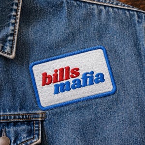 May include: A rectangular embroidered patch attached to a blue denim jacket. The patch features a white background with a blue border and the text "bills mafia" in red and blue embroidered lettering, with "bills" in red and "mafia" in blue.