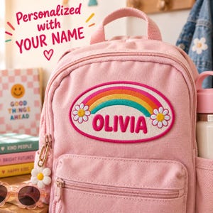 May include: A light pink backpack featuring a large oval patch on the front. The patch displays a colourful rainbow, two white daisies, and the name OLIVIA in bold pink letters. A white daisy charm hangs from the zipper pull of the front pocket.