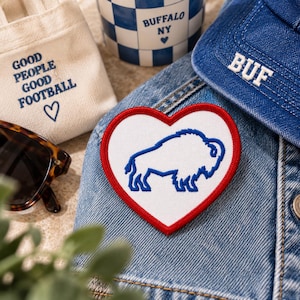 Könnte beinhalten: Ein herzförmiger Aufnäher mit weißem Hintergrund, rotem Rand und einer blauen gestickten Bison-Kontur. Der Aufnäher liegt auf einer blauen Jeansjacke neben einer cremefarbenen Stofftasche mit dem Text GOOD PEOPLE GOOD FOOTBALL und einem Herz.