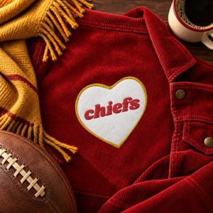 Pode incluir: Um casaco de veludo cotelê vermelho com um emblema branco em forma de coração com o texto vermelho "chiefs" bordado no centro. O casaco é acompanhado por um cachecol amarelo-mostarda com franjas e uma bola de futebol americano castanha.