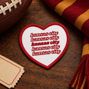 Pode incluir: Um emblema bordado em forma de coração com fundo branco e borda vermelha. O texto 'kansas city' é repetido cinco vezes em vermelho, empilhado verticalmente numa fonte estilizada no centro do emblema, criando um efeito visual em camadas.