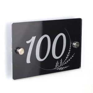 Peut inclure: Plaque de numéro de maison en acrylique noir avec inscription argentée et un motif floral. Le numéro 100 est affiché sur la plaque.