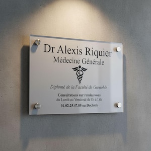May include: A clear acrylic sign with silver fixings, mounted on a grey wall. The sign displays black text: "Dr Alexis Riquier, Médecine Générale," a medical symbol, and other details. The sign is for a medical practice.