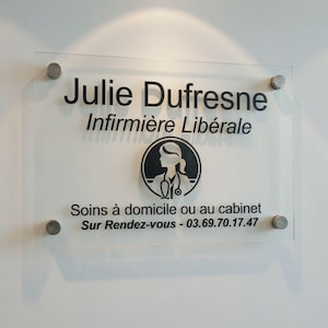 Puede incluir: Un letrero de acrílico transparente con el nombre "Julie Dufresne" y el título "Infirmière Libérale" en letras negras. Un logotipo circular de una enfermera está centrado debajo del nombre. El letrero está montado con separadores plateados.