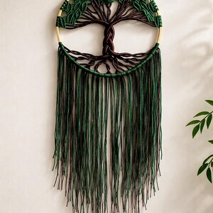 Op de afbeelding: Een macramé wandhanger met een boomontwerp. De boom heeft een bruine stam en takken, met groene bladeren. Het cirkelvormige frame is goudkleurig. Lange strengen groen en bruin macramé hangen eronder.