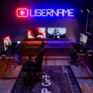 Puede incluir: Un letrero de neón rojo y azul con la palabra "USERNAME" y un botón de reproducción. El letrero está montado en una pared sobre una configuración de juego con dos monitores de computadora, un teclado y una silla de juego.
