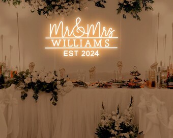 May include: A white neon sign with the text "Mr & Mrs WILLIAMS EST 2024" on a white wall. The sign is illuminated with a warm white glow. The sign is decorated with white flowers.