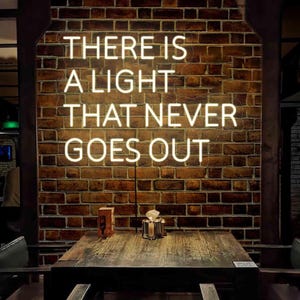 Könnte beinhalten: Ein weißes Neon-Schild mit dem Text "There is a light that never goes out" an einer Backsteinwand montiert. Das Schild hängt über einem Holztisch mit zwei Stühlen.