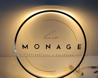 Könnte beinhalten: Ein rundes, weißes, beleuchtetes Schild mit schwarzem Rand. Das Schild trägt die Aufschrift "hair MONAGE STYLED BY VALL X RNLS STYLES" in goldenen Buchstaben.