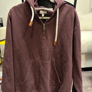 May include: A maroon hoodie with a half-zip closure and a kangaroo pocket. The hood features white drawstrings with brown leather-like accents. The hoodie is hanging on a white hanger with the word "LOVE" printed on it.