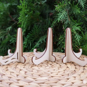 May include: Three wooden display stands with a light wood finish and dark brown edges. The stands have a curved base and a tall, slender back, perfect for displaying small items. The stands are arranged on a woven surface against a backdrop of green foliage.