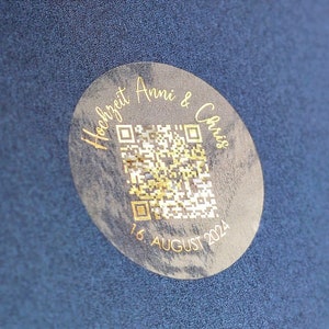 May include: A round, clear sticker with a gold design. The sticker says "Hochzeit Anni & Chris" and "16. AUGUST 2024" and features a QR code.
