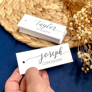 Può includere: Etichette rettangolari bianche con nomi calligrafici neri "Taylor MAZZIOTTI" e "joseph CONCEPCION". Le etichette hanno un piccolo foro in alto a sinistra. Le etichette sono su un tappetino intrecciato e una superficie blu scuro.