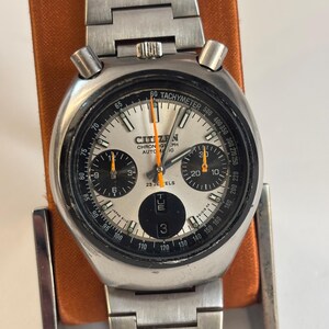 May include: Vintage Citizen chronograph automatic watch with a silver dial, black subdials, and a stainless steel band. The watch features a tachymeter bezel and orange hands. The watch is displayed on an orange stand.