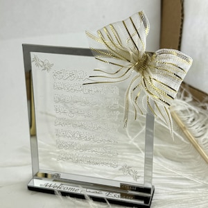 May include: A clear glass plaque with an etched Arabic inscription and a gold and white bow on top. The plaque has a black base with the words "Welcome عصا ESSAM" engraved on it.