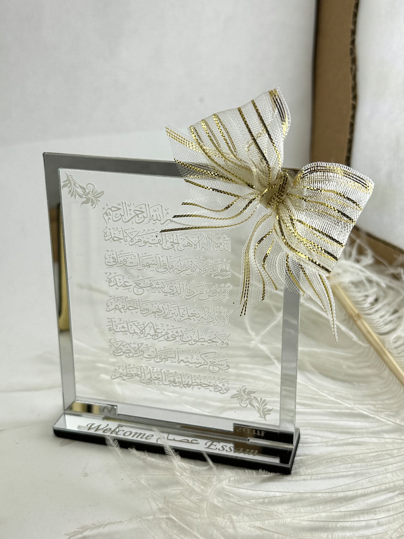 May include: A clear glass plaque with an etched Arabic inscription and a gold and white bow on top. The plaque has a black base with the words "Welcome عصا ESSAM" engraved on it.