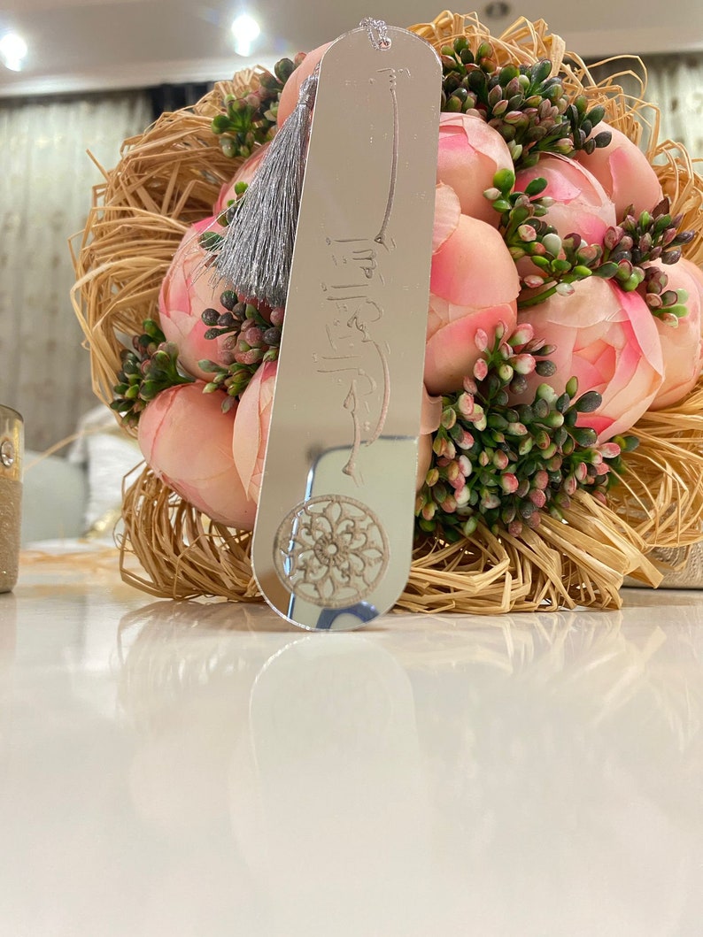 May include: A silver bookmark with a tassel and an engraved design. The bookmark is resting on a bouquet of pink flowers.