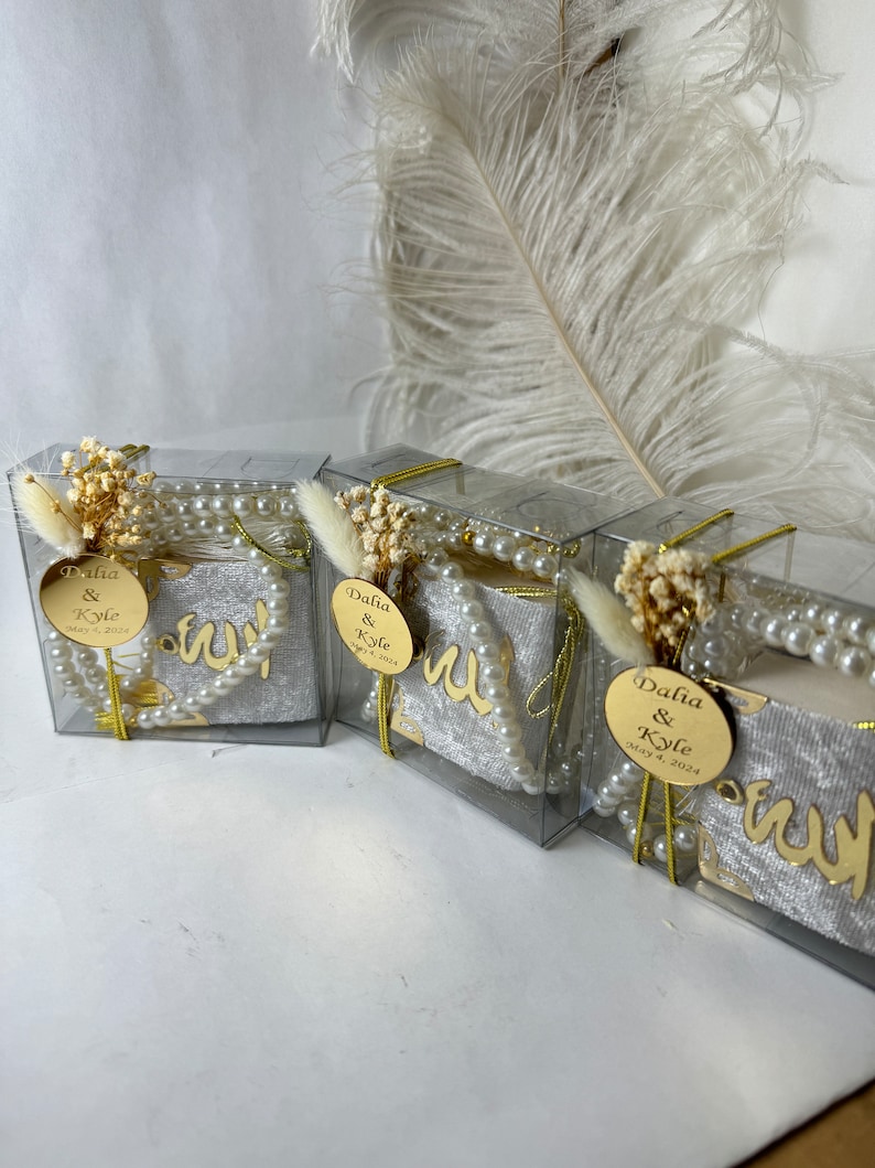May include: Three clear boxes with white velvet fabric inside. Each box has a gold circle with the text "Dalia & Kyle May 4, 2024" and a string of white pearls.