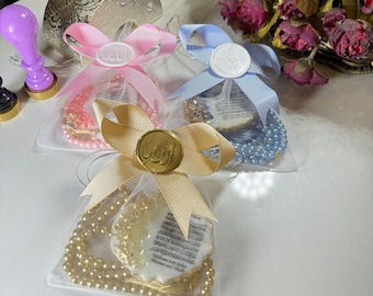 May include: Three small gift bags with white tulle and gold ribbon. Each bag contains a white cookie with gold leaf and Arabic script, and a pearl necklace. The bags are decorated with a pink, blue, and gold bow with a wax seal.
