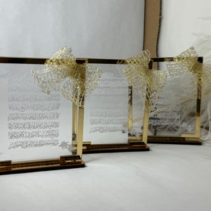 May include: Three gold-framed glass plaques with Arabic calligraphy and a gold ribbon on each. The plaques are on a white surface.