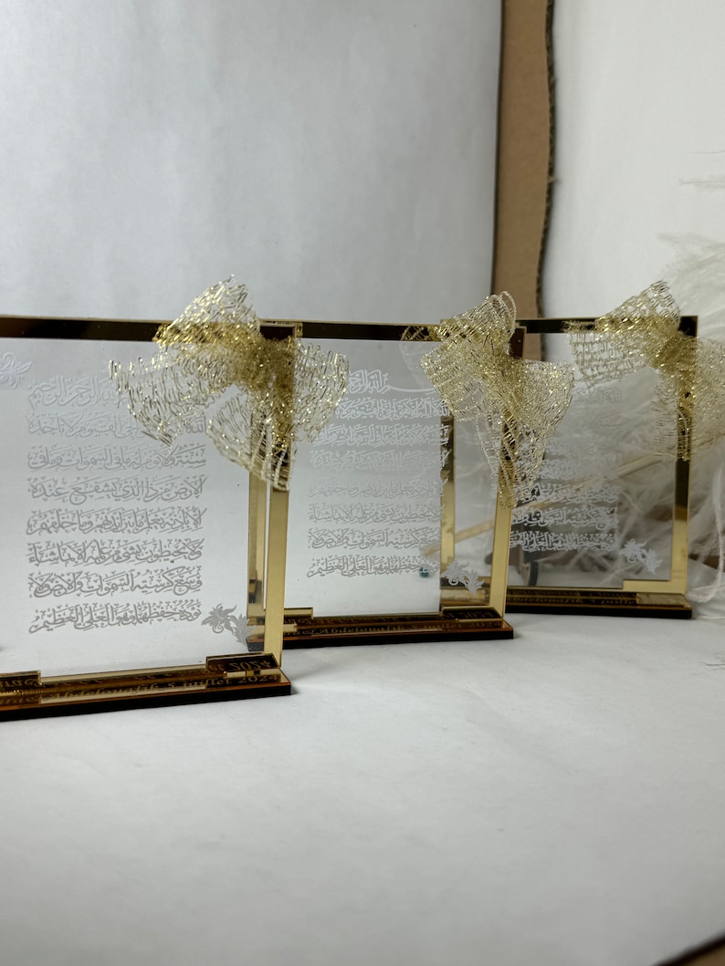 May include: Three gold-framed glass plaques with Arabic calligraphy and a gold ribbon on each. The plaques are on a white surface.