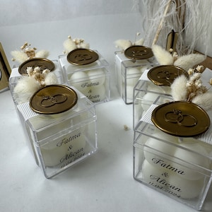 May include: Clear plastic boxes filled with white candles, each box has a gold wax seal with a ring design and a label with the text "Fatma & Alican 11.04.2024".