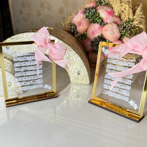 May include: Two gold-framed glass plaques with white calligraphy and pink ribbons. The plaques are on a white surface with a bouquet of pink flowers in the background.