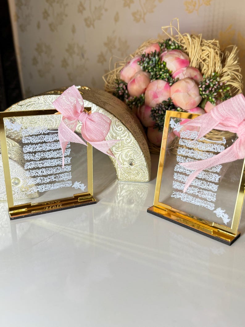 May include: Two gold-framed glass plaques with white calligraphy and pink ribbons. The plaques are on a white surface with a bouquet of pink flowers in the background.