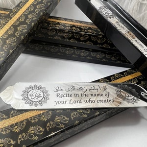 May include: A silver bookmark with a black background and gold accents. The bookmark features an Arabic inscription that reads "Recite in the name of your Lord who created." The bookmark is in a black box with a gold pattern.