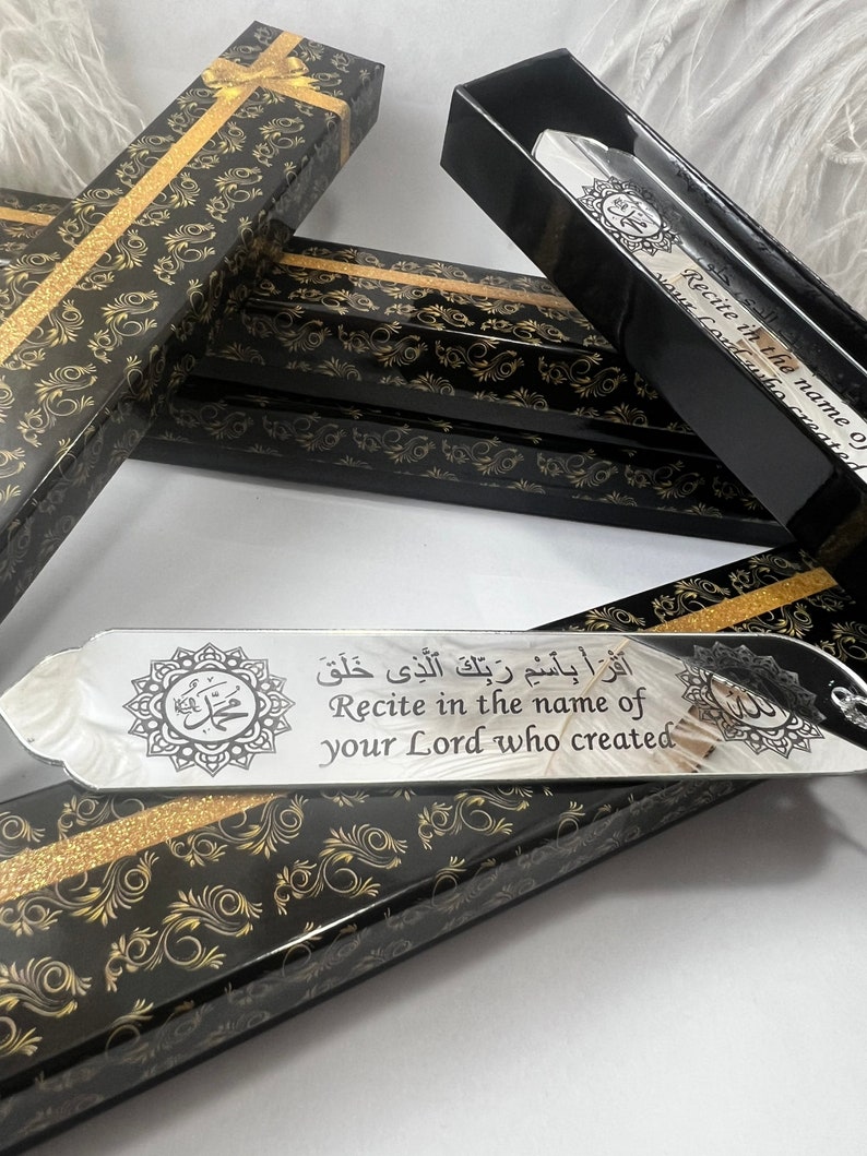 May include: A silver bookmark with a black background and gold accents. The bookmark features an Arabic inscription that reads "Recite in the name of your Lord who created." The bookmark is in a black box with a gold pattern.