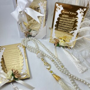 May include: A white prayer bead necklace with a gold charm and tassel, a gold framed card with Arabic text and the words "Ayet el Kursi" and a white gift box with a white bow.