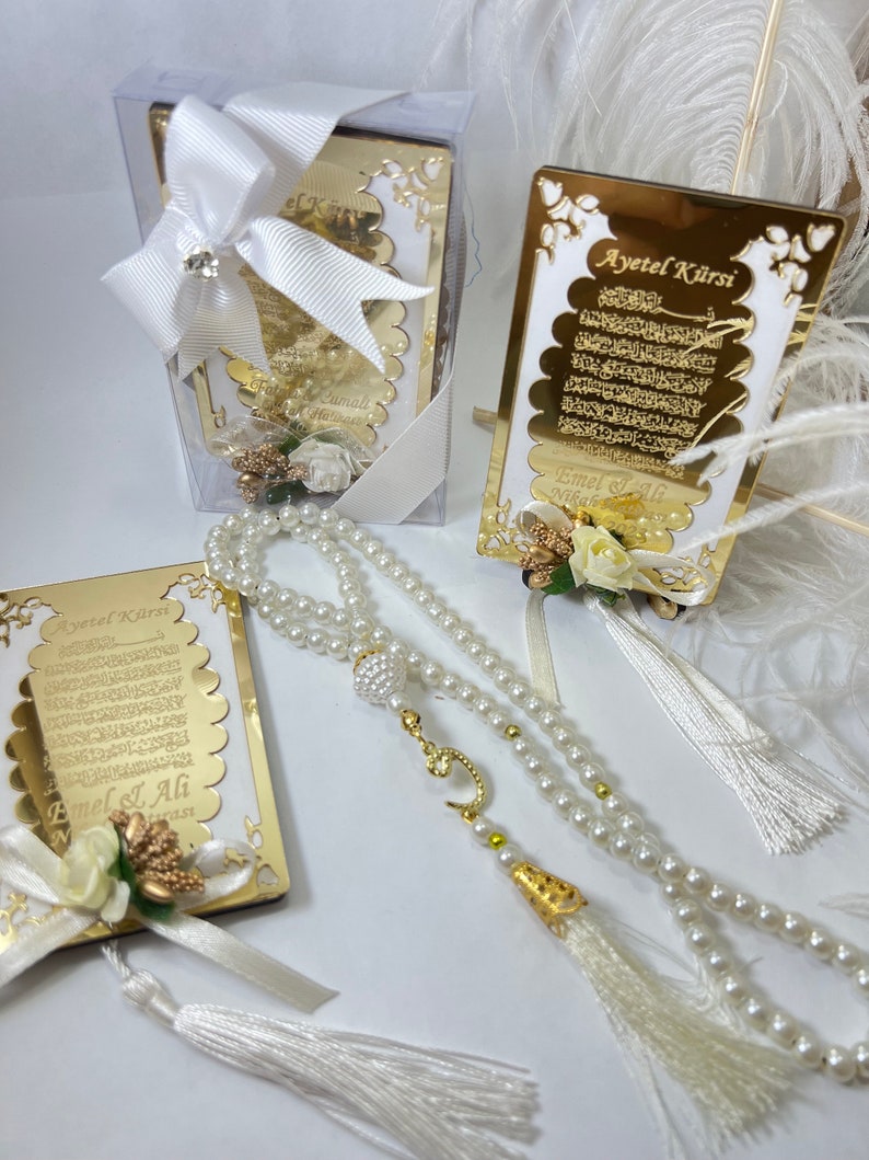 May include: A white prayer bead necklace with a gold charm and tassel, a gold framed card with Arabic text and the words "Ayet el Kursi" and a white gift box with a white bow.