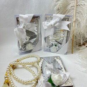 May include: Two silver boxes with white satin bows and white flowers. The boxes contain small white prayer beads. A larger set of white prayer beads with a gold tassel is in the foreground.