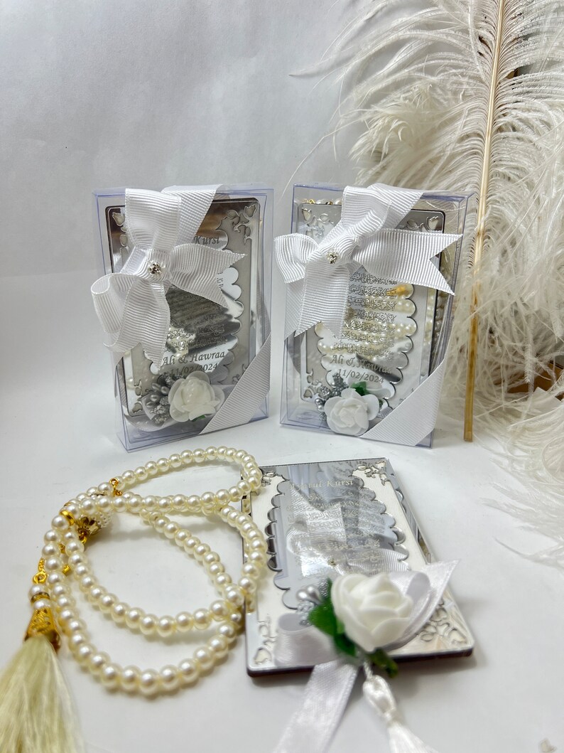 May include: Two silver boxes with white satin bows and white flowers. The boxes contain small white prayer beads. A larger set of white prayer beads with a gold tassel is in the foreground.